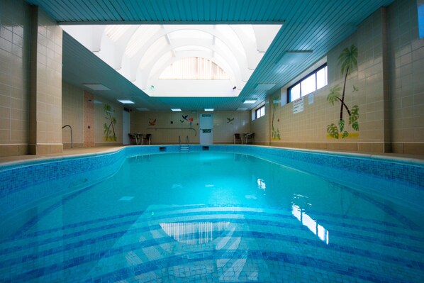 An indoor swimming pool with a tiled floor and walls, a high ceiling with skylights, and murals of palm trees and birds on the walls. - Best Western York House Hotel (Eastbourne)