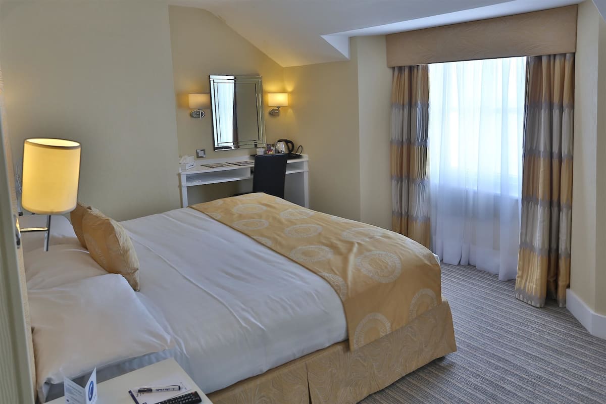Een hotelkamer met een bed, nachtkastje, lamp, bureau, spiegel en gordijnen.