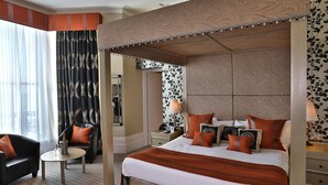 A hotel room with a four-poster bed, a sitting area with a chair and table, and a large window with sheer curtains.