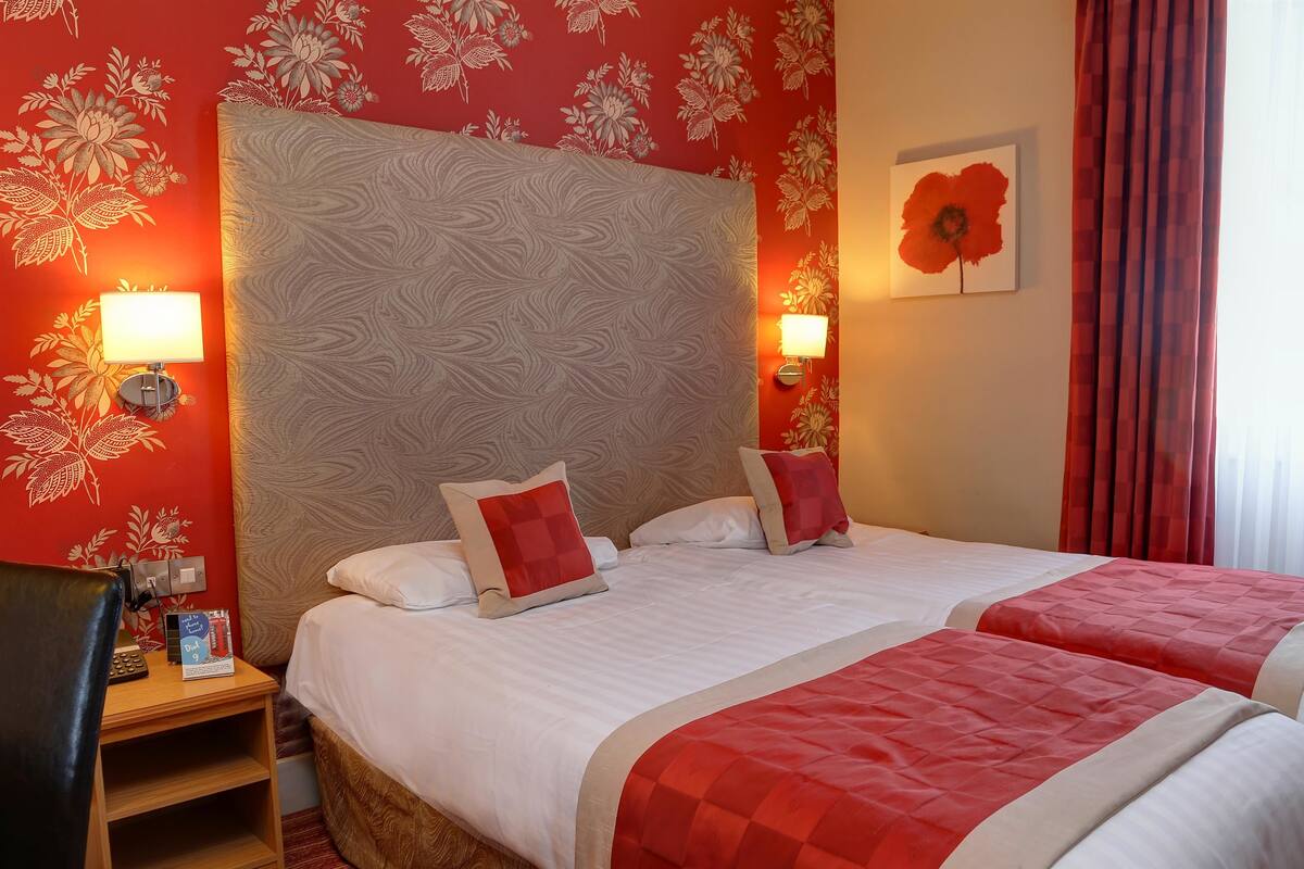 Een hotelkamer met twee bedden, bloemenbehang, een groot hoofdeinde en een schilderij van een rode bloem aan de muur.