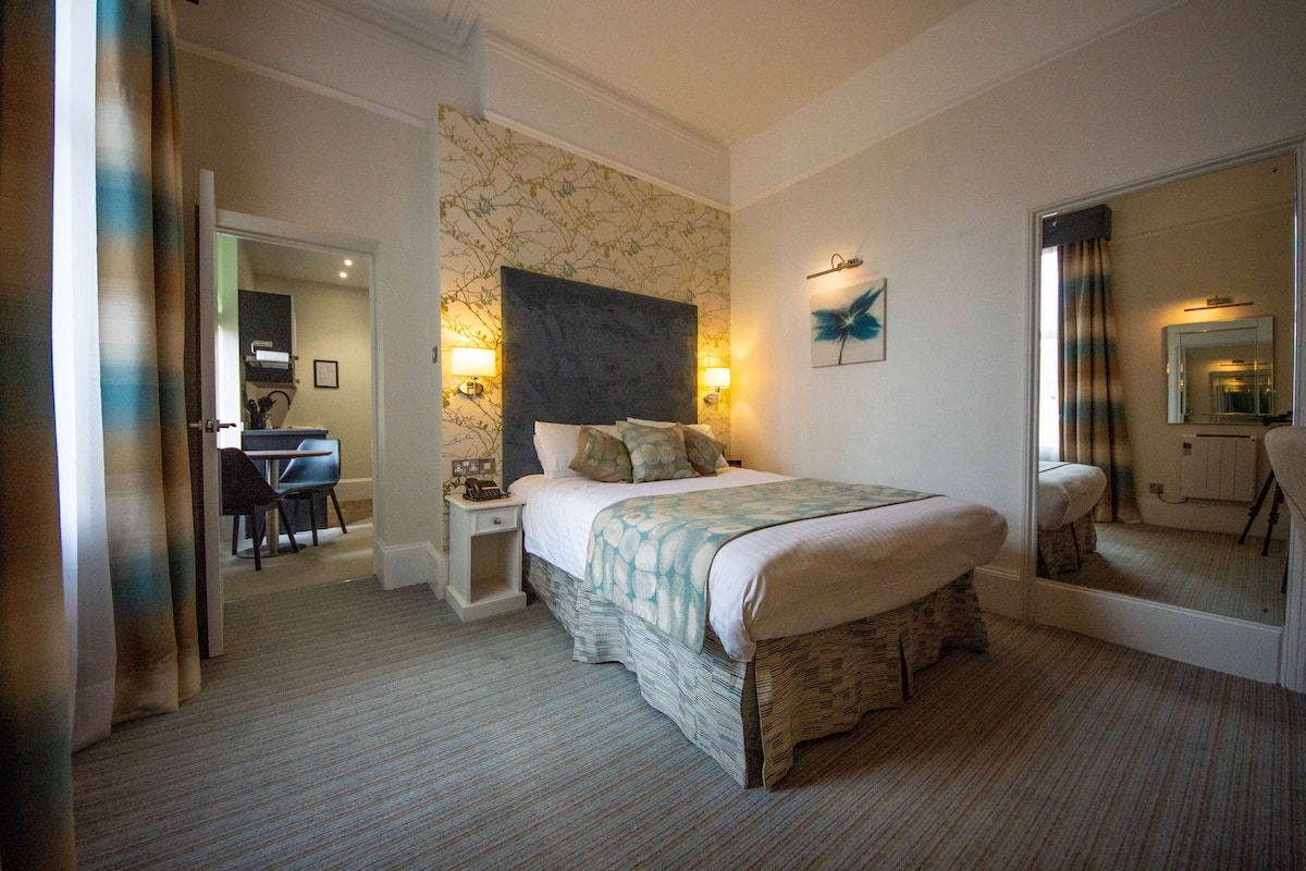 Une chambre d’hôtel avec un grand lit, un bureau avec une chaise, un miroir et un mur orné d’un motif floral.