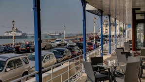 A seaside café with outdoor seating overlooking a parking area and a pier.