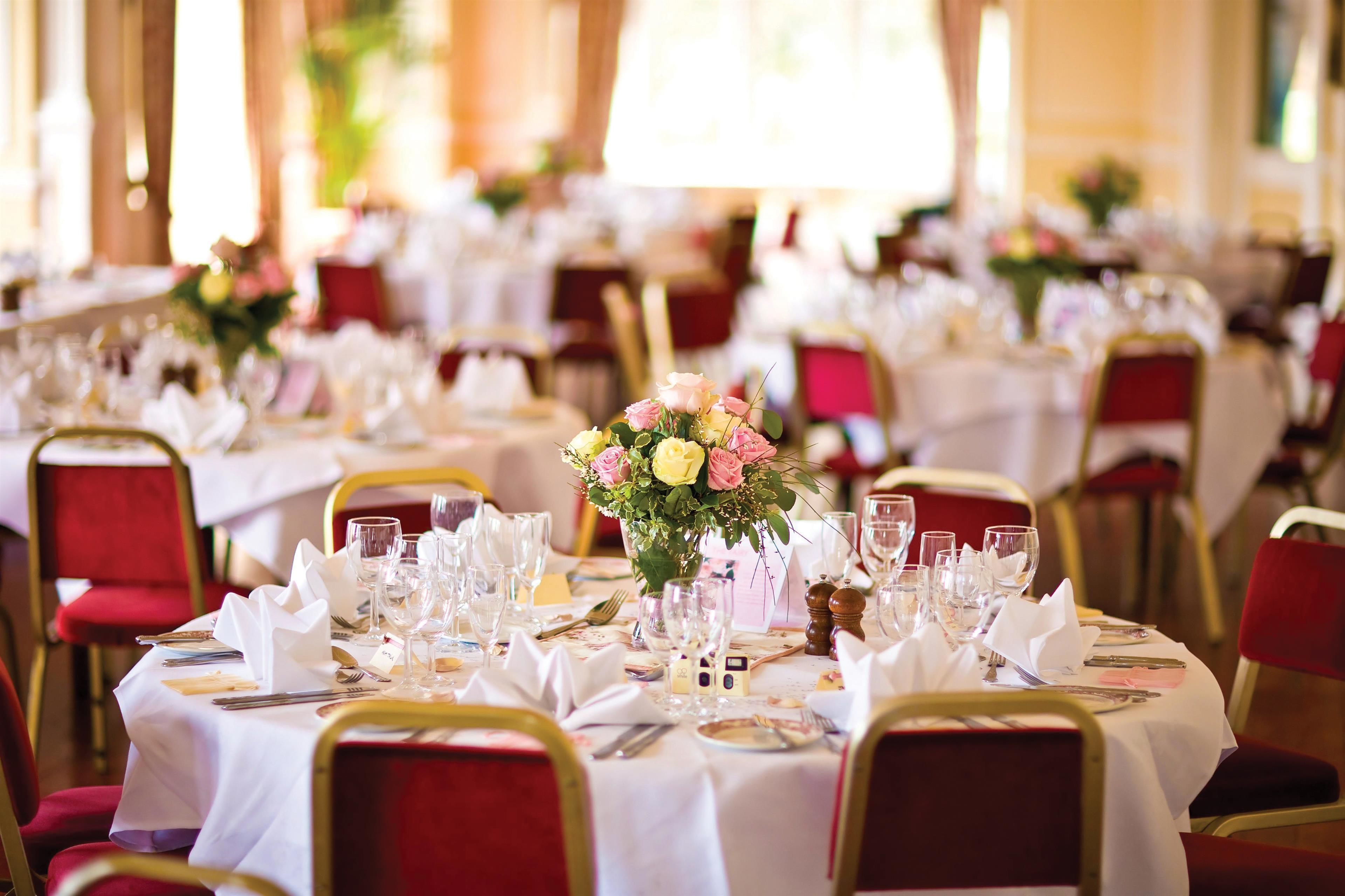 A banquet hall with round tables set for dining, each with white tablecloths, red chairs, and floral centerpieces.
