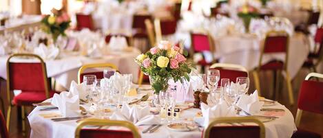 A banquet hall with round tables set for dining, each with white tablecloths, red chairs, and floral centerpieces.