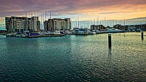 A marina with boats docked, buildings in the background, and calm water.