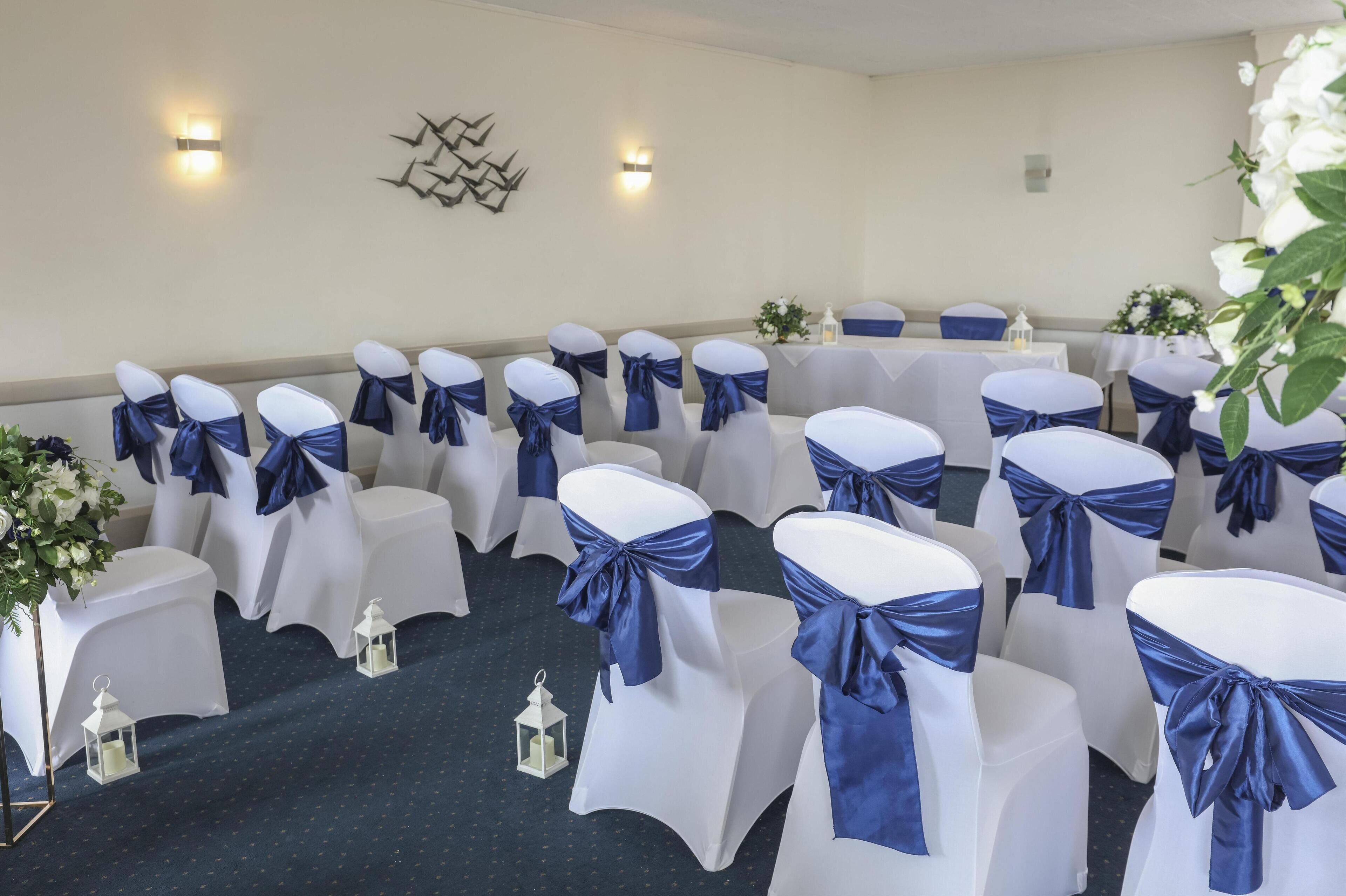 Una sala conferenze con file di sedie bianche decorate con nastri blu, una moquette a motivi e lampade fissate al muro.