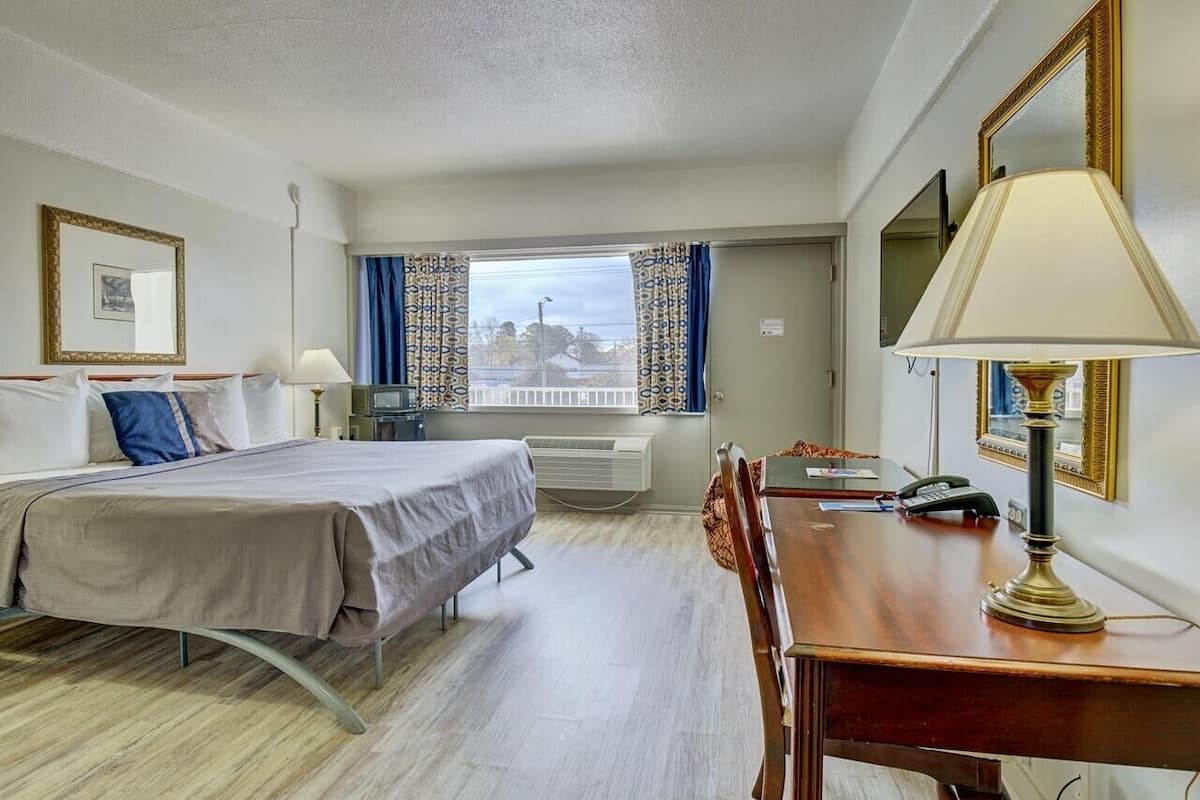 A hotel room with a bed, desk, and chair, a window with curtains, and a mirror on the wall.