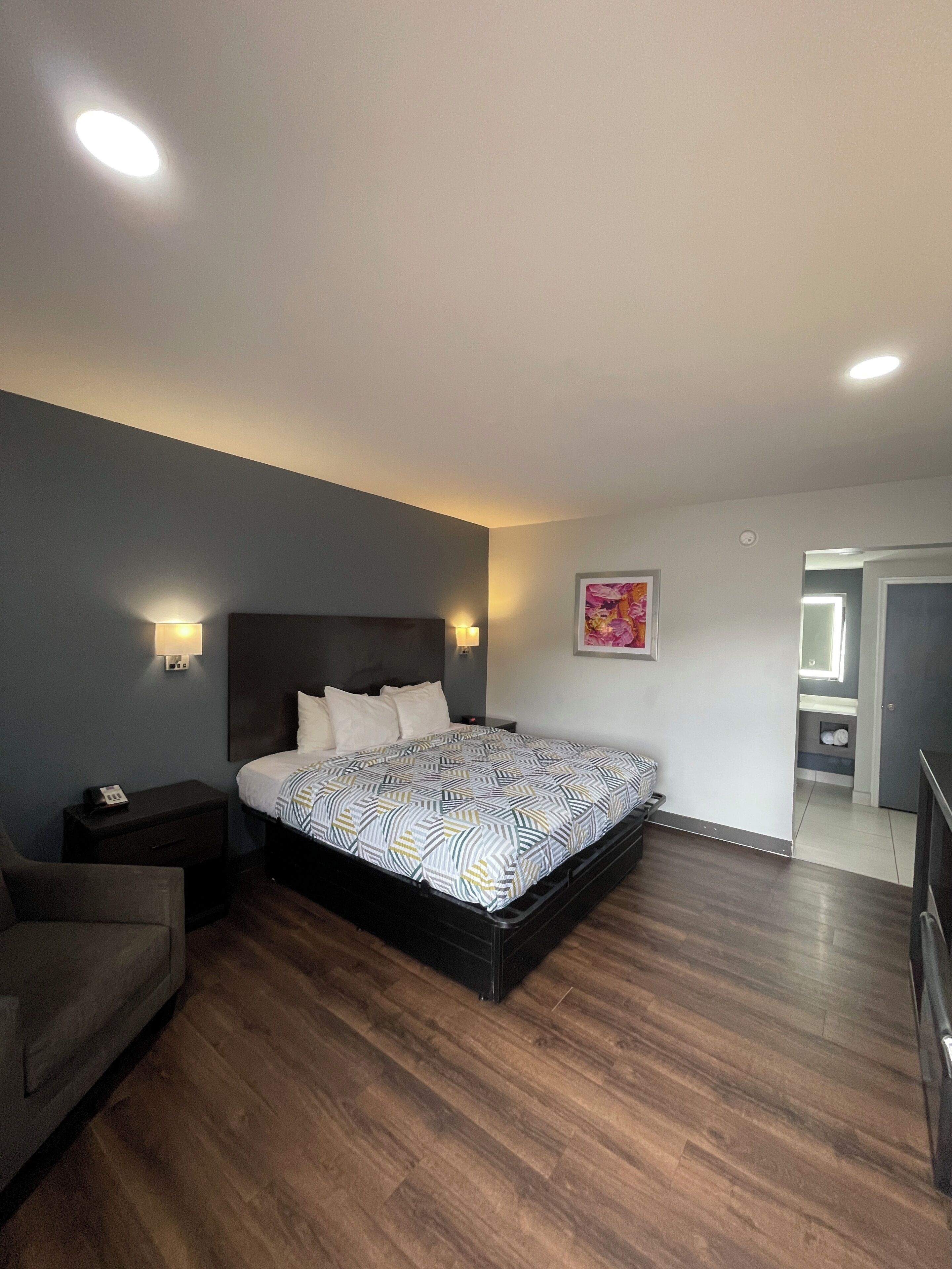 A bedroom with a bed, bedside tables, a nightstand, a chair, and a bathroom visible through an open door.
