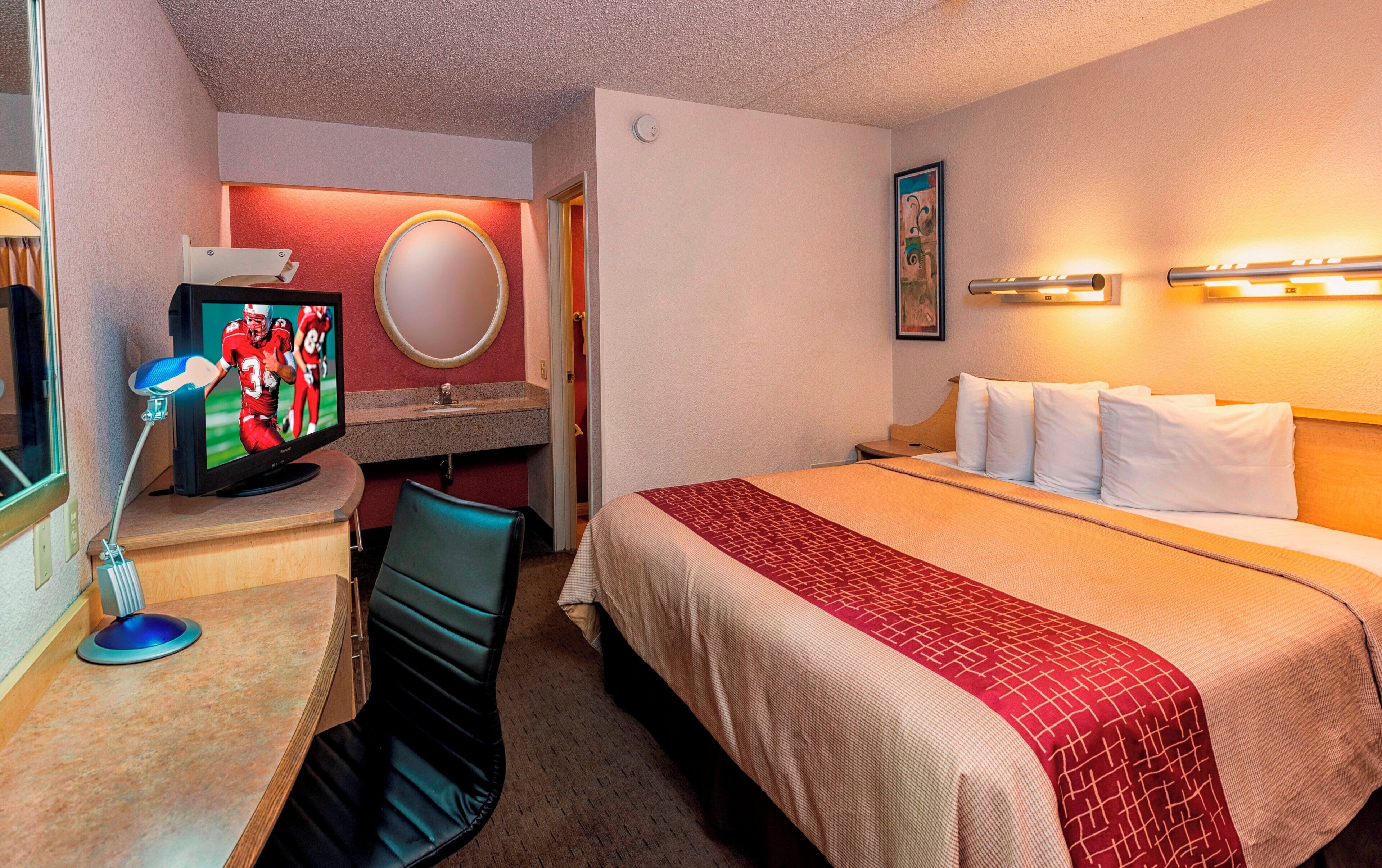 Habitación de hotel con cama, escritorio con televisor, silla, espejo y lámpara de pared.