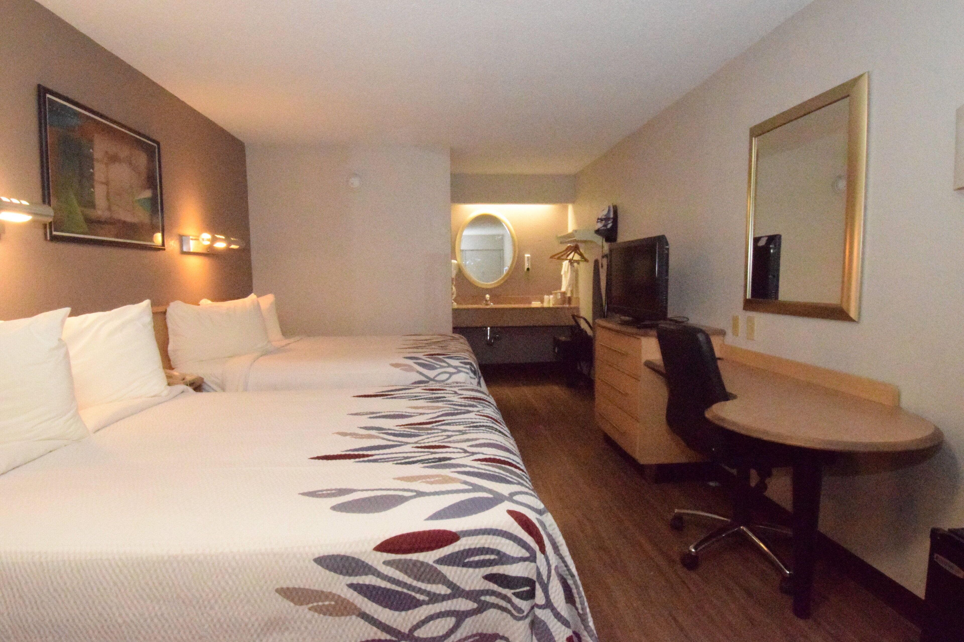 Une chambre d’hôtel avec deux lits, un bureau, une chaise, une télévision et un miroir. On aperçoit également une salle de bain au fond.