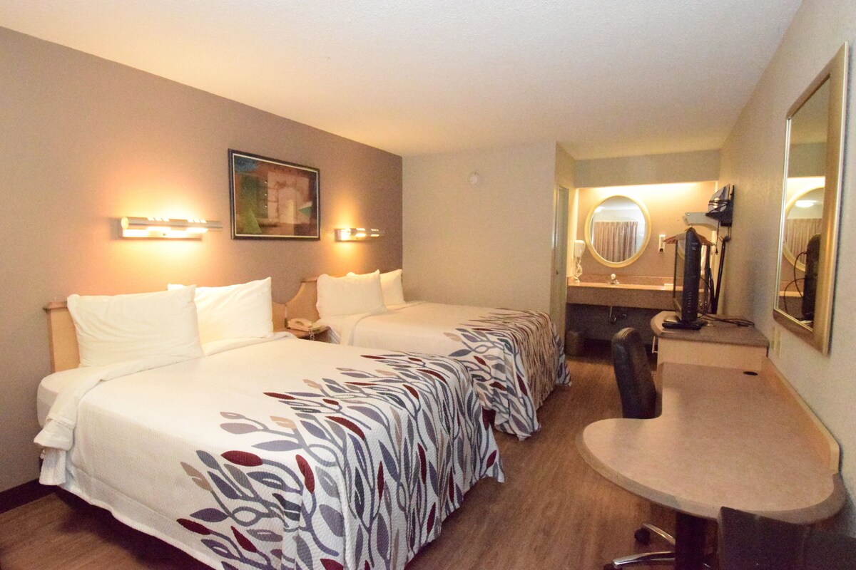 A hotel room with two beds, a desk, a chair, a mirror, and a bathroom with a round mirror.