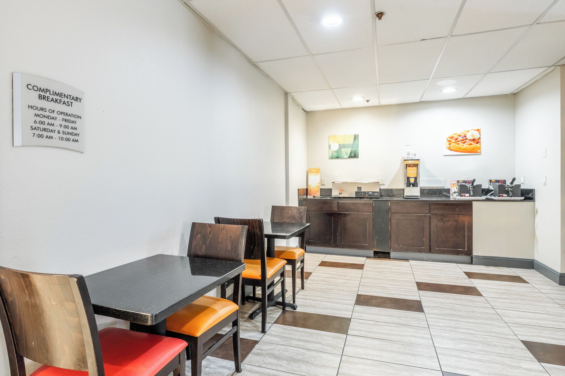 A small cafe with a table and chairs, a counter with a coffee machine, and a sign indicating complimentary breakfast hours.