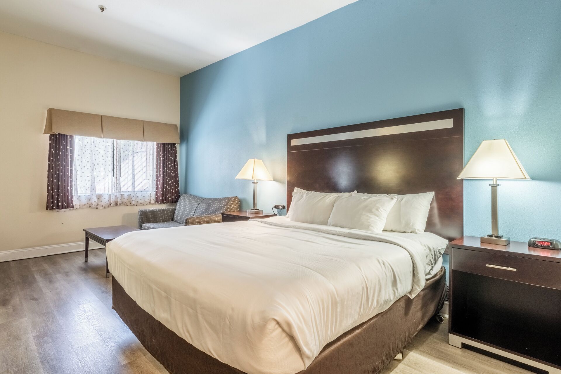 Una habitación de hotel con una cama grande, dos mesitas de noche con lámparas, un pequeño sofá y un ventanal con cortinas.