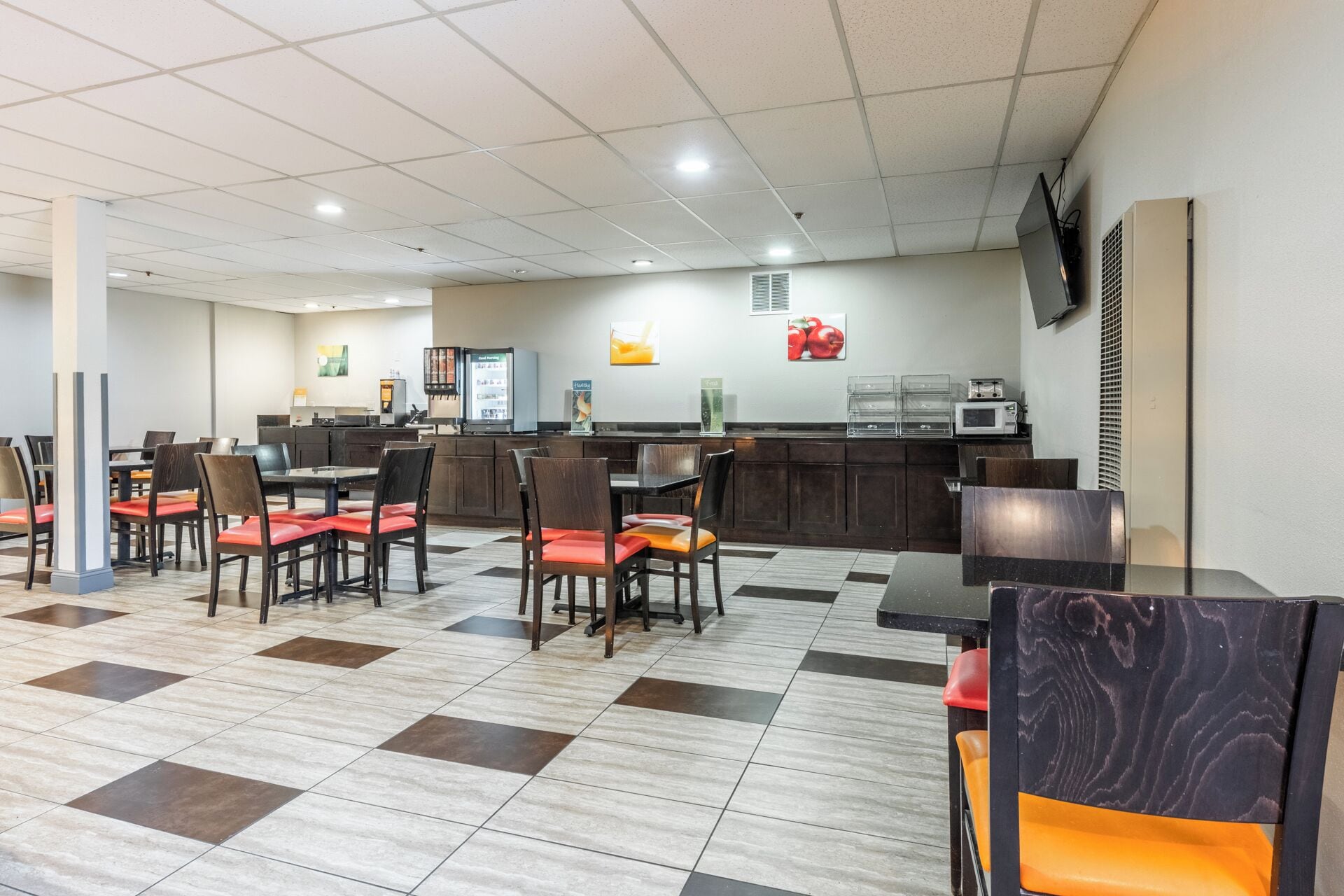 A clean, well-lit restaurant area with a checkered floor, wooden tables and chairs, a self-service beverage station, and a microwave on the counter.