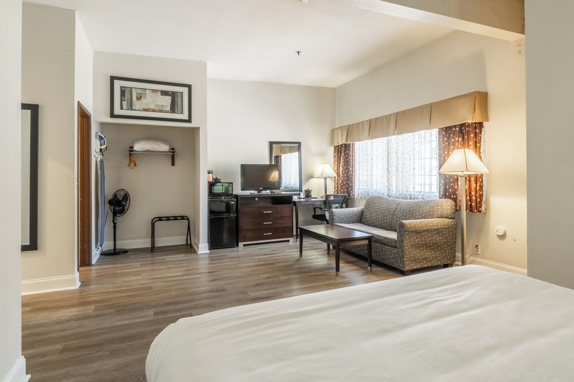 A hotel room with a bed, a desk with a computer, a chair, a sofa, a small table, a fan, and a window with curtains.