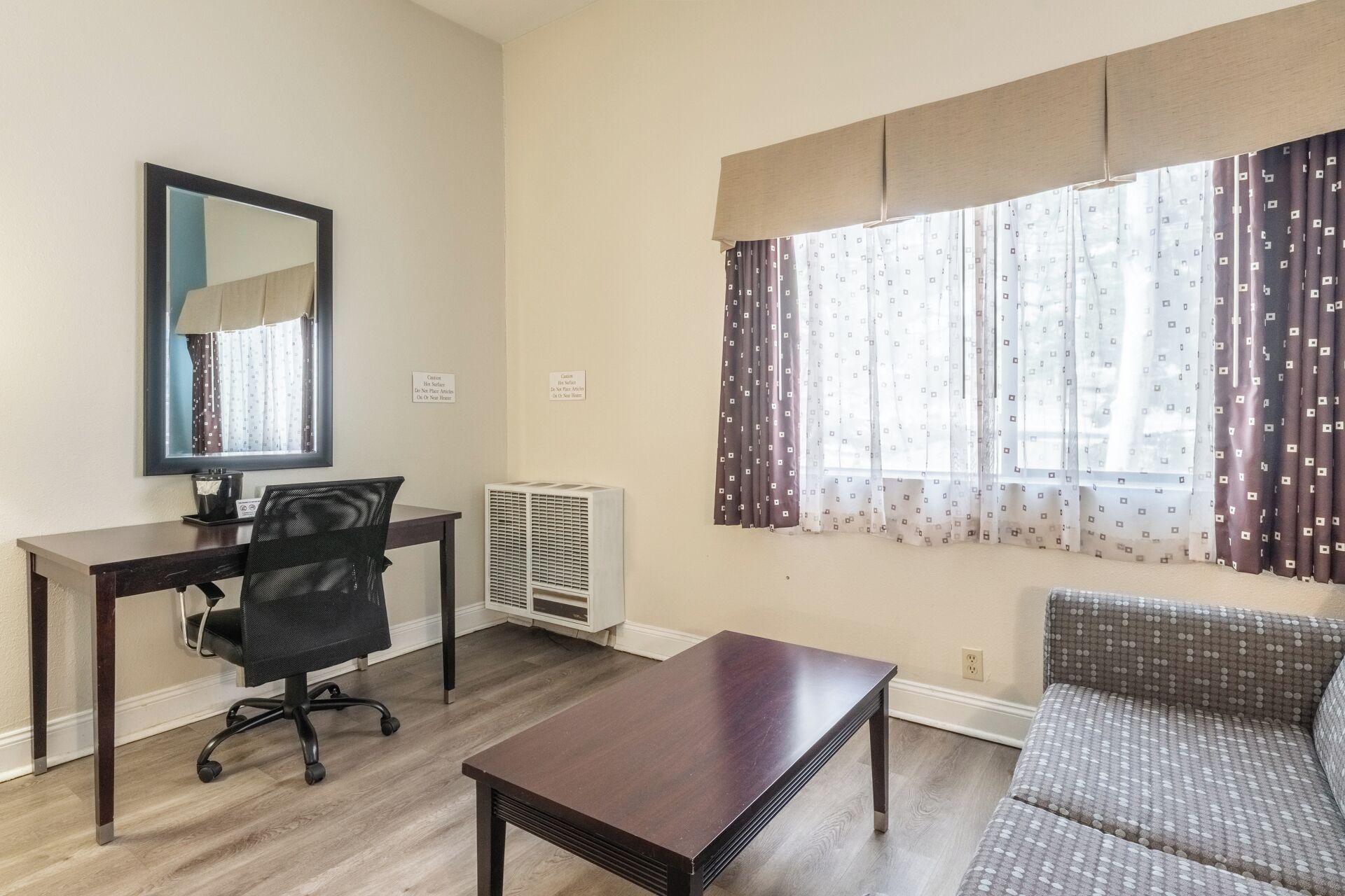 Una sala de estar con una mesa de madera oscura, una silla de oficina negra, un sofá, una ventana con cortinas estampadas y un espejo montado en la pared.