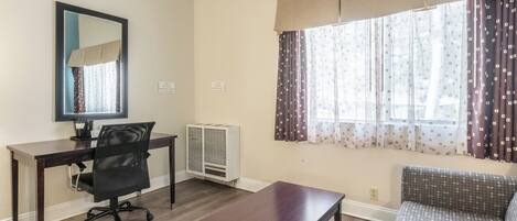 Una sala de estar con una mesa de madera oscura, una silla de oficina negra, un sofá, una ventana con cortinas estampadas y un espejo montado en la pared.