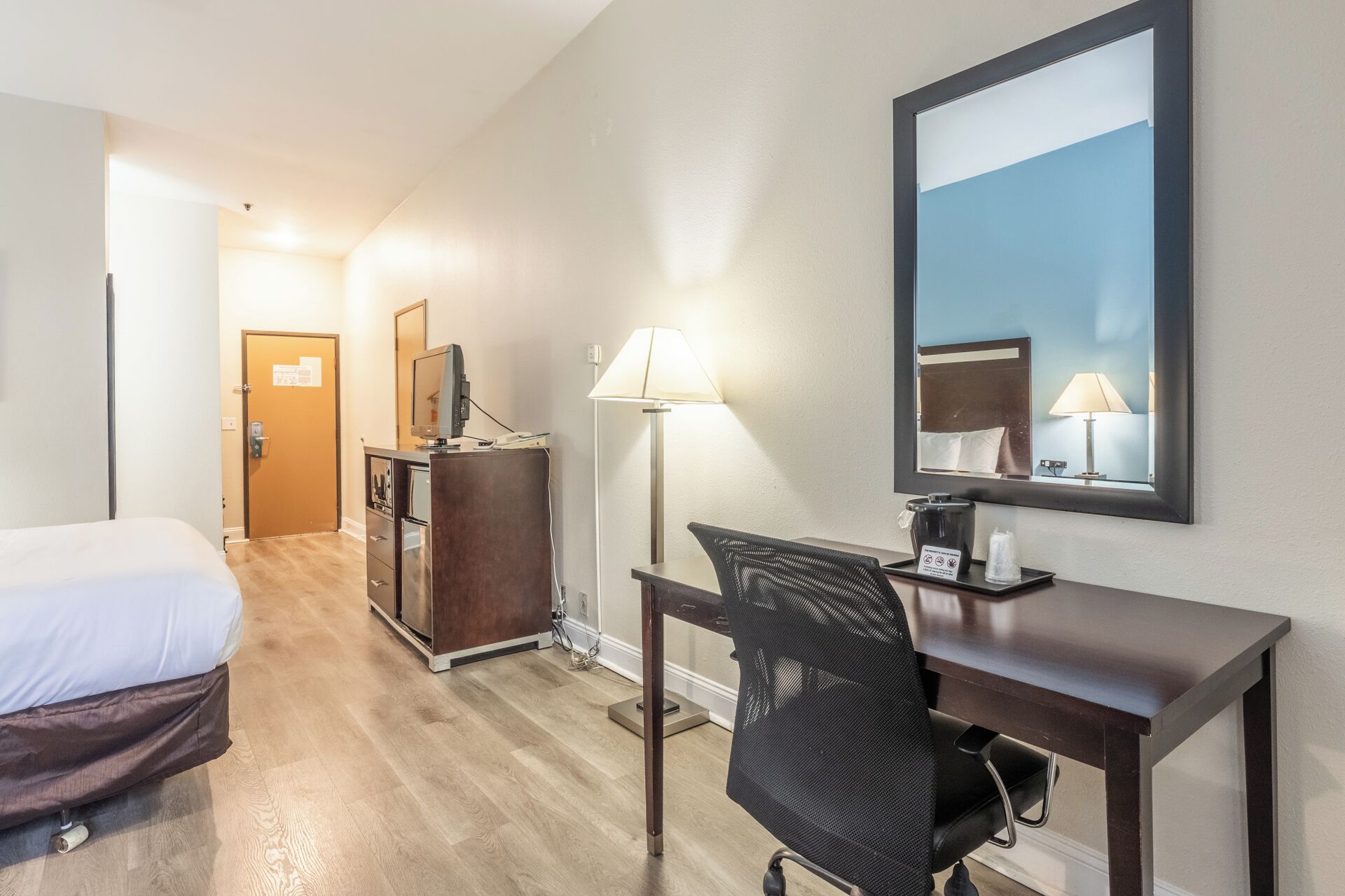 A hotel room with a bed, a desk with a chair, a dresser, a mirror, and a lamp.