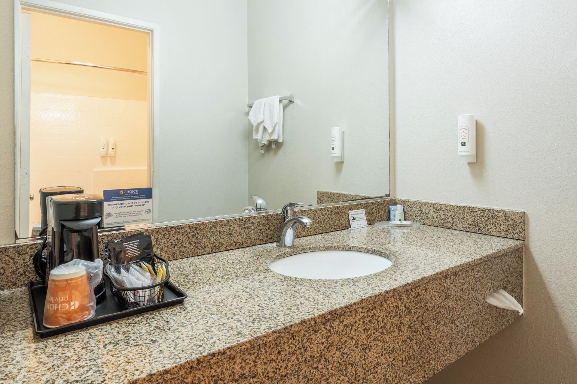Casa de banho de hotel com bancada de granito, uma pia, um espelho, uma máquina de café e um porta-toalhas.