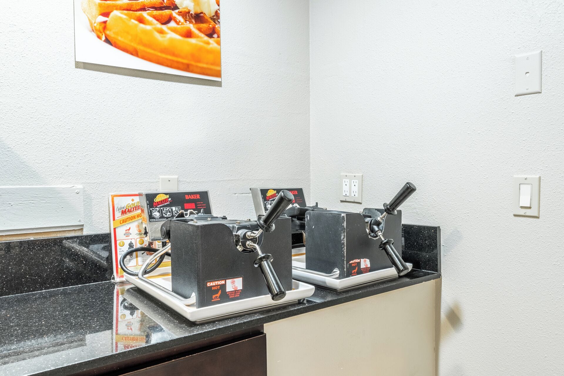 Een keukenhoek met twee wafelijzers op een zwart aanrecht, een waarschuwingsbord en een wandcontactdoos.