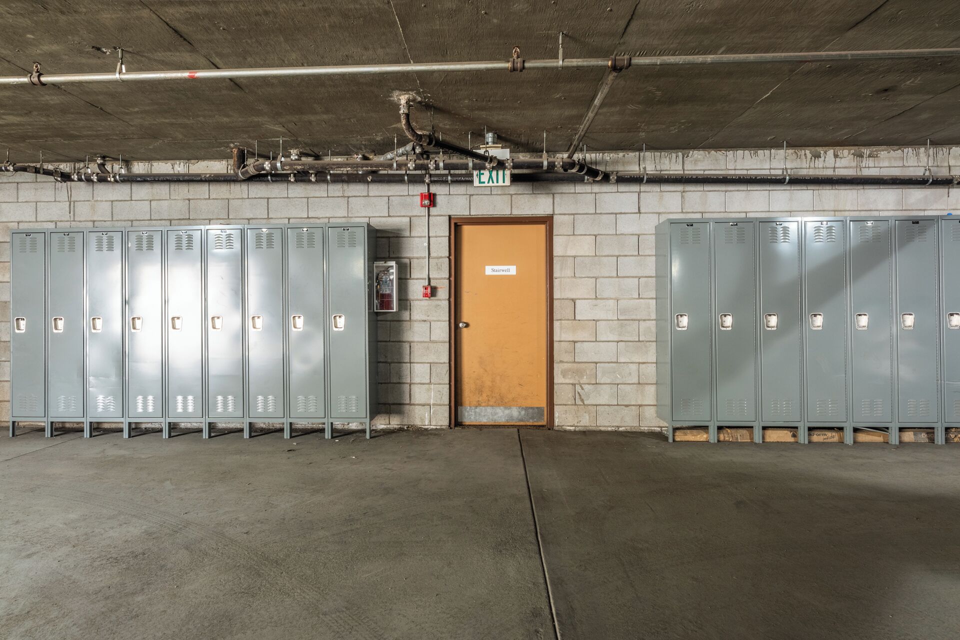 Een praktische kleedruimte met meerdere metalen kastjes, een houten deur met de tekst 'Trappenhuis' en zichtbare leidingen en brandveiligheidsmaterialen.