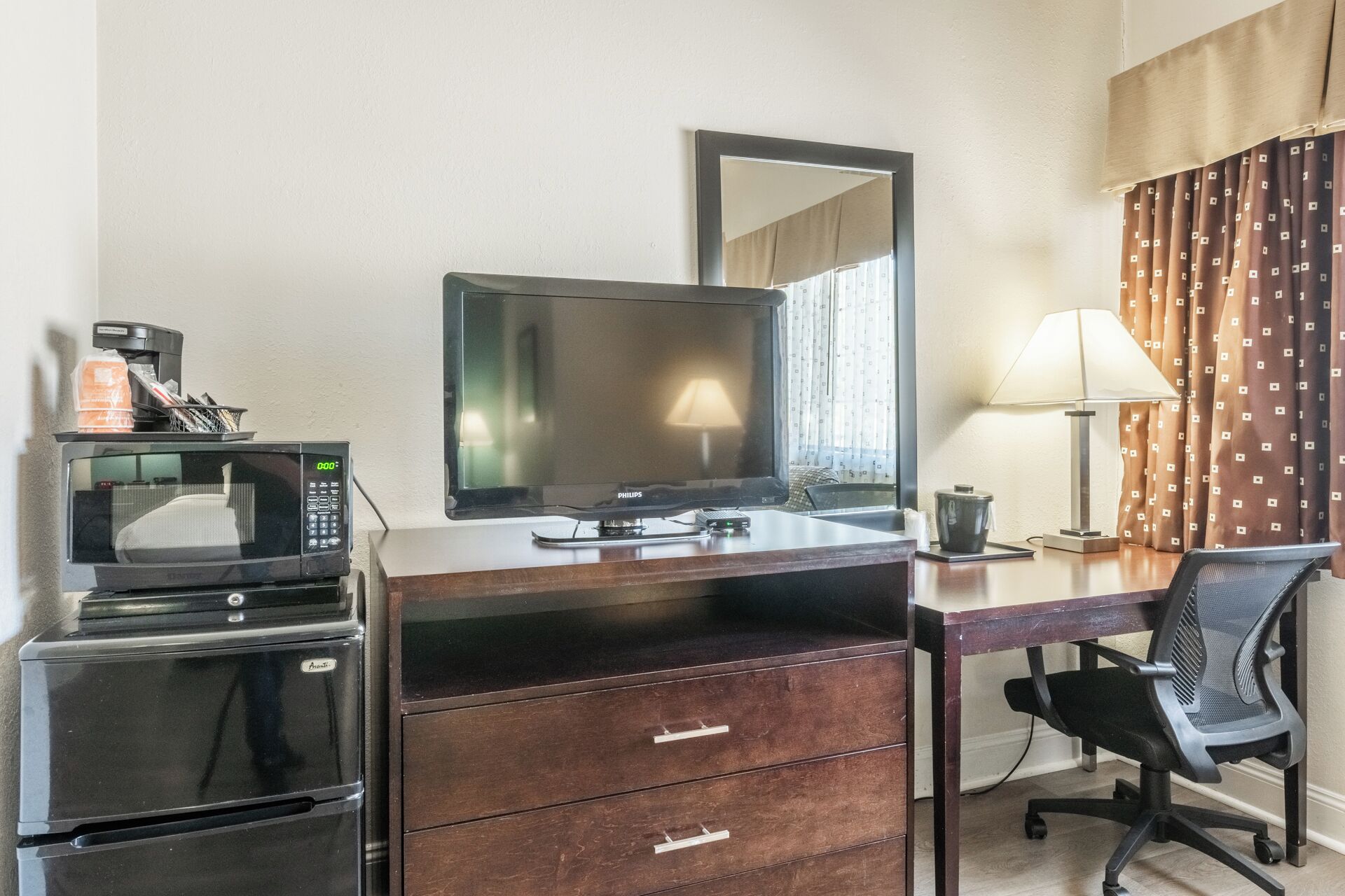 A hotel room with a desk, a flat-screen TV, a microwave, a coffee maker, a lamp, and a chair.