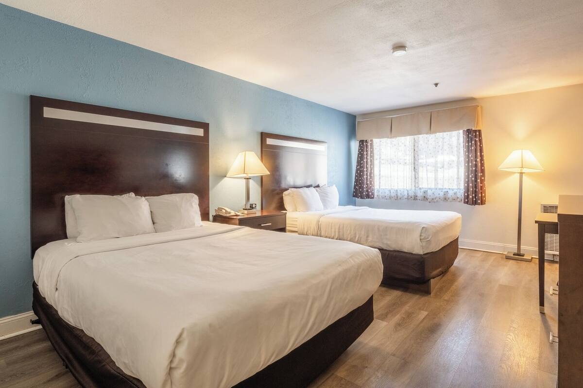 A hotel room with two beds, a nightstand with a lamp, a window with curtains, and a ceiling light.
