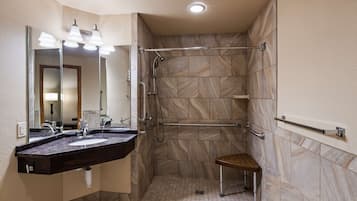 Un baño moderno con un espejo grande, lavamanos dobles, ducha con barra de apoyo y un banco pequeño.