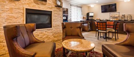 A modern living room with leather armchairs, a round coffee table, a flat-screen TV mounted on a brick wall, and a kitchen area with a coffee machine and breakfast bar.