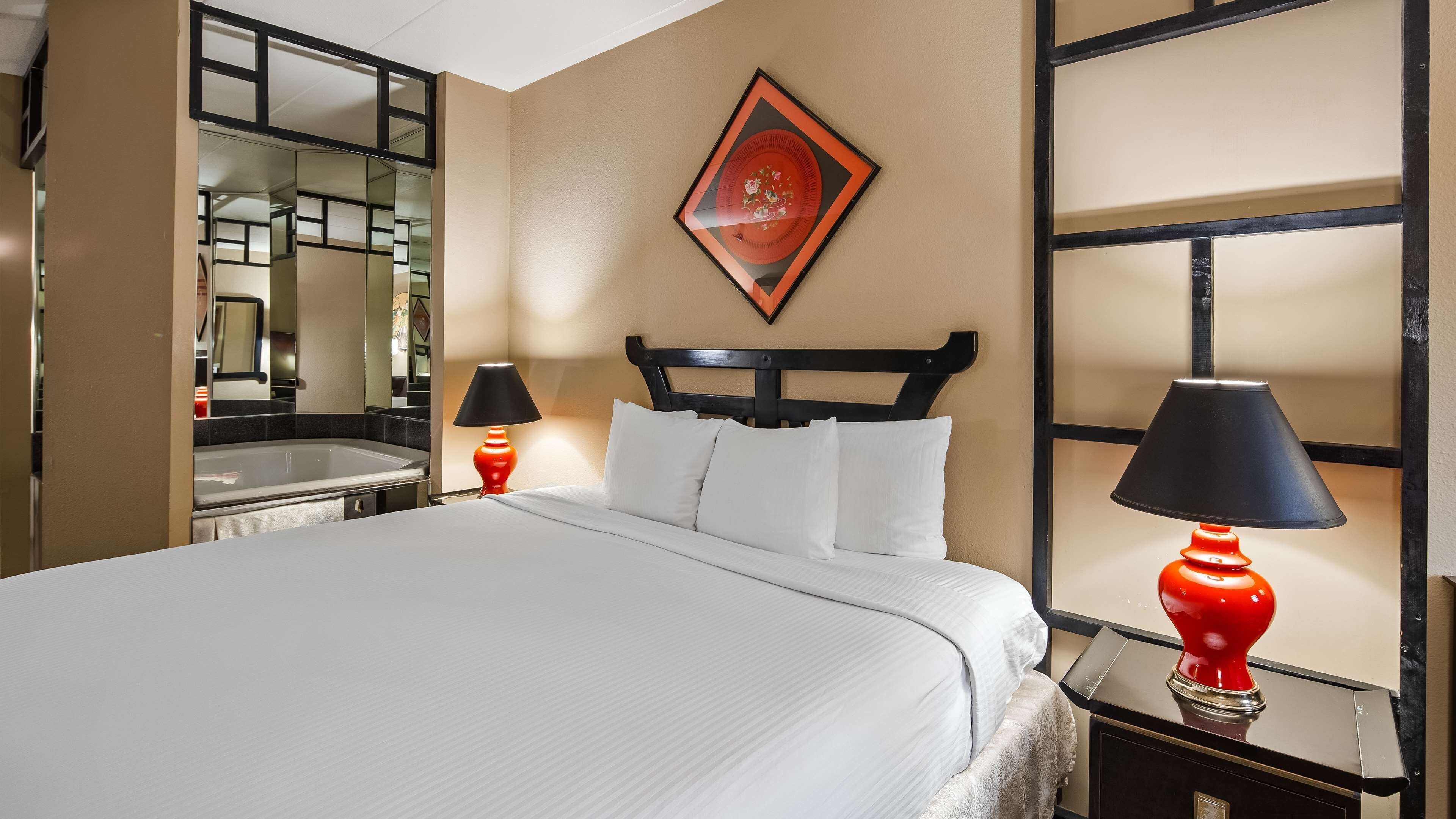 Une chambre d’hôtel avec un lit, deux lampes de chevet, un miroir, un objet décoratif rouge accroché au mur et un tableau encadré au-dessus du lit.