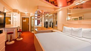Una habitación de hotel moderna con una cama grande, una zona de estar con un sillón rojo, un tocador con espejo y un baño pequeño.