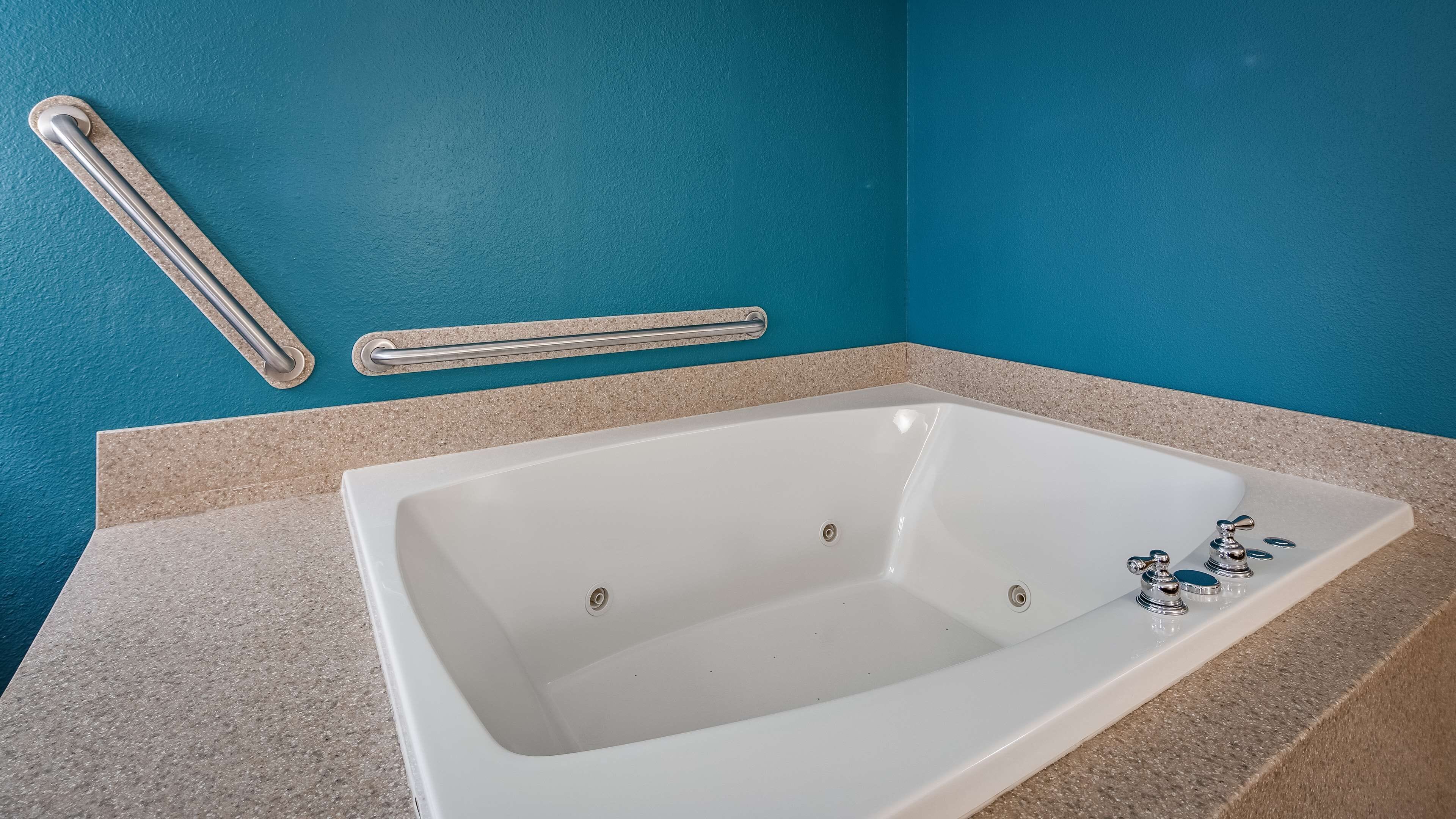 Une baignoire blanche avec un seul robinet et une douchette à main, une barre d’appui et un porte-serviettes, le tout adossé à un mur de couleur turquoise.