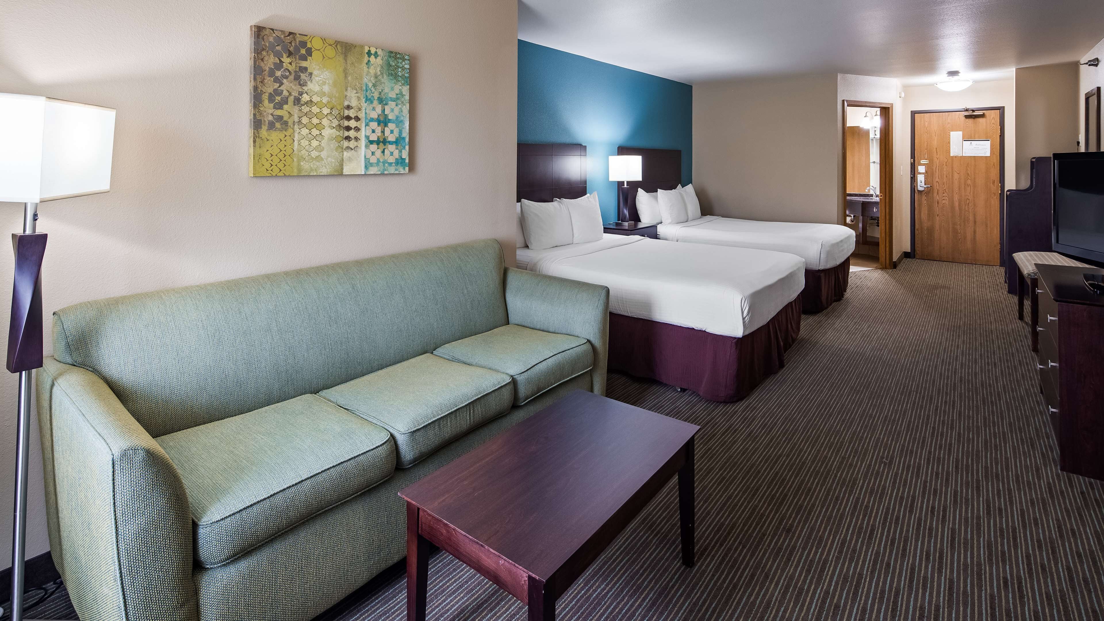 A hotel room with a green sofa, a wooden coffee table, two beds, a TV, and a painting on the wall.