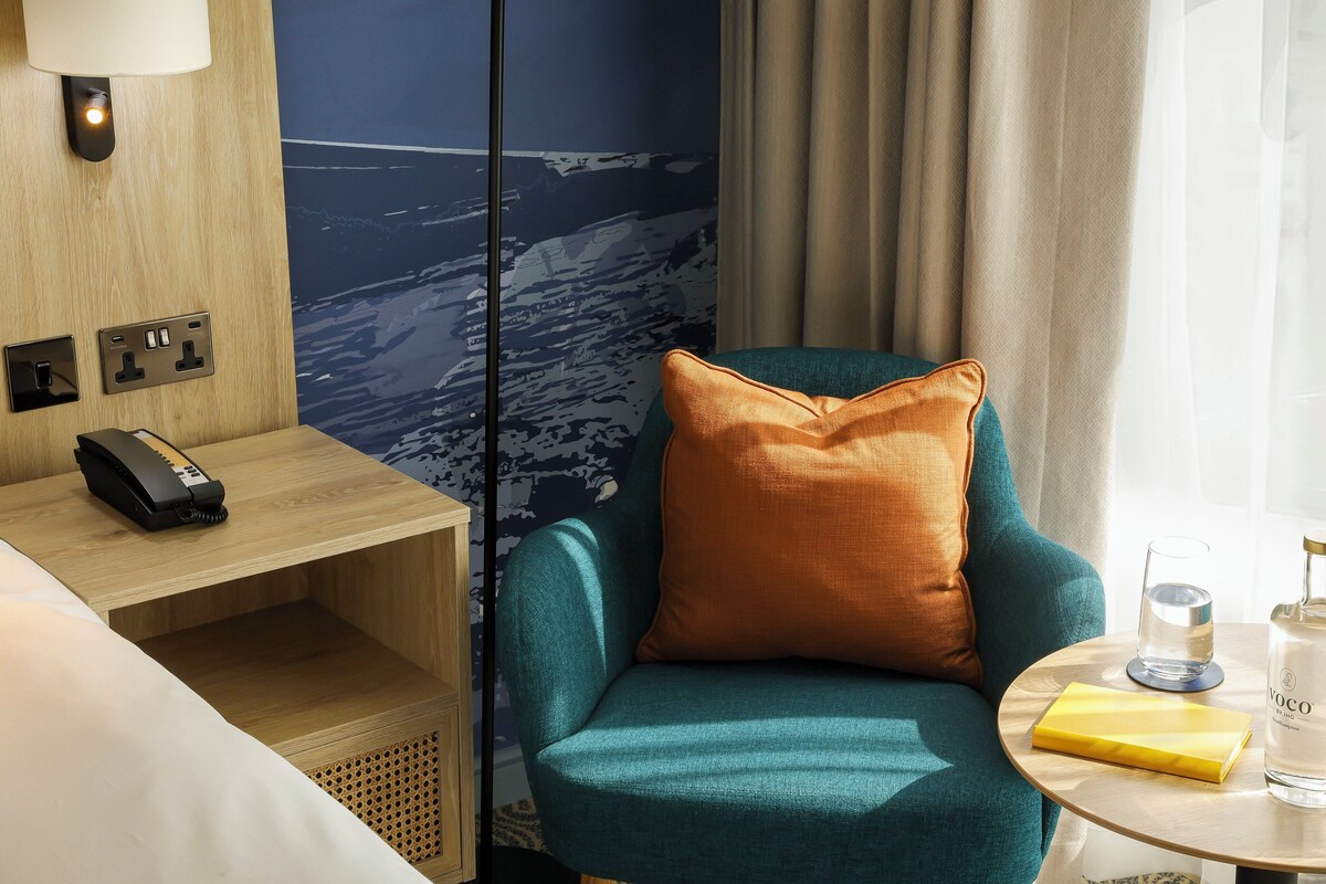 Habitació d'hotel amb una poltrona color aguamarina, una tauleta de nit de fusta amb un telèfon, un got d'aigua i una ampolla de licor sobre una taula petita.