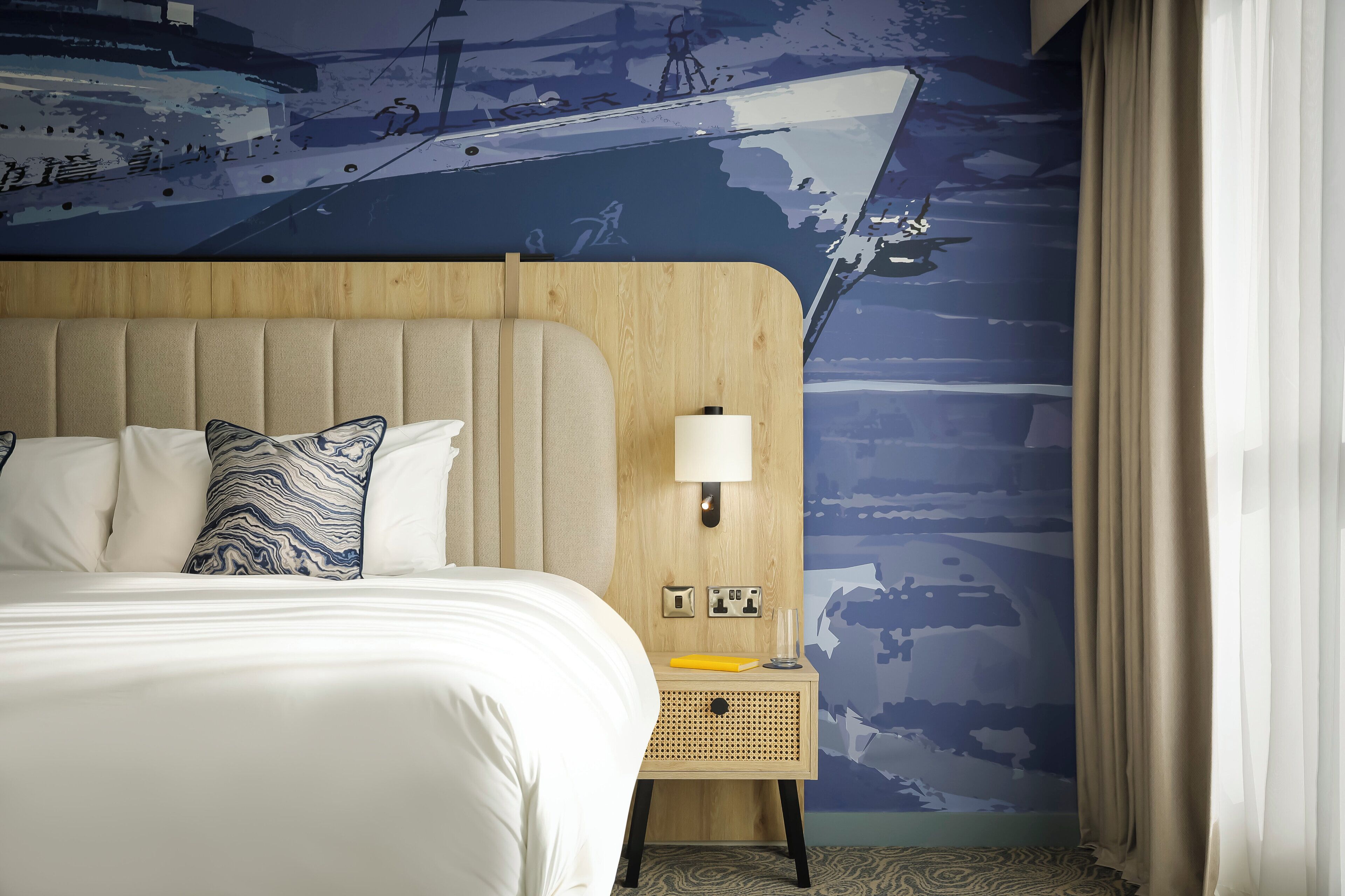 A hotel room with a large bed, a wooden headboard, a nightstand with a lamp, and a wall mural.