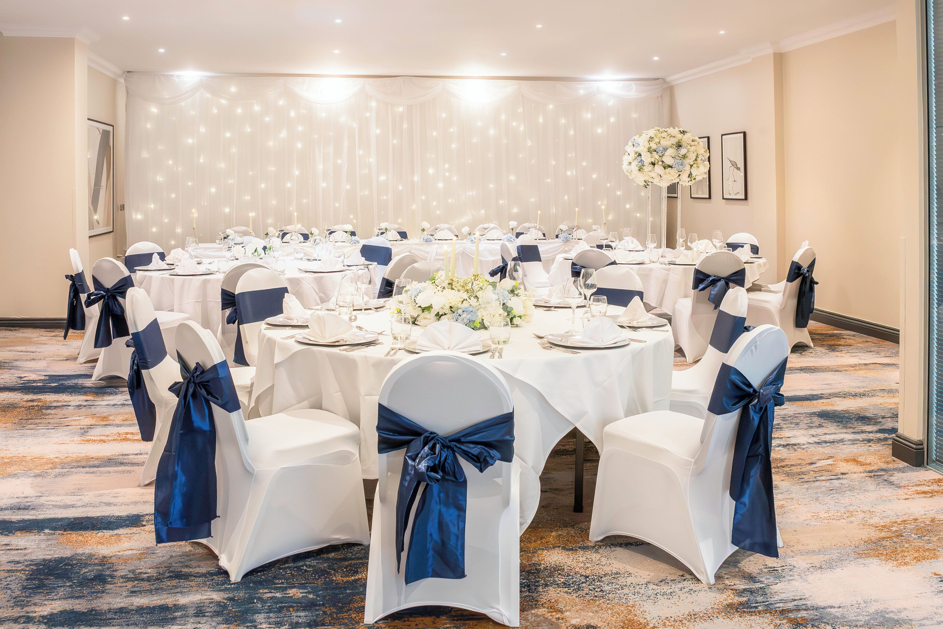 A banquet hall with round tables, white chairs with blue sashes, and a floral centerpiece.