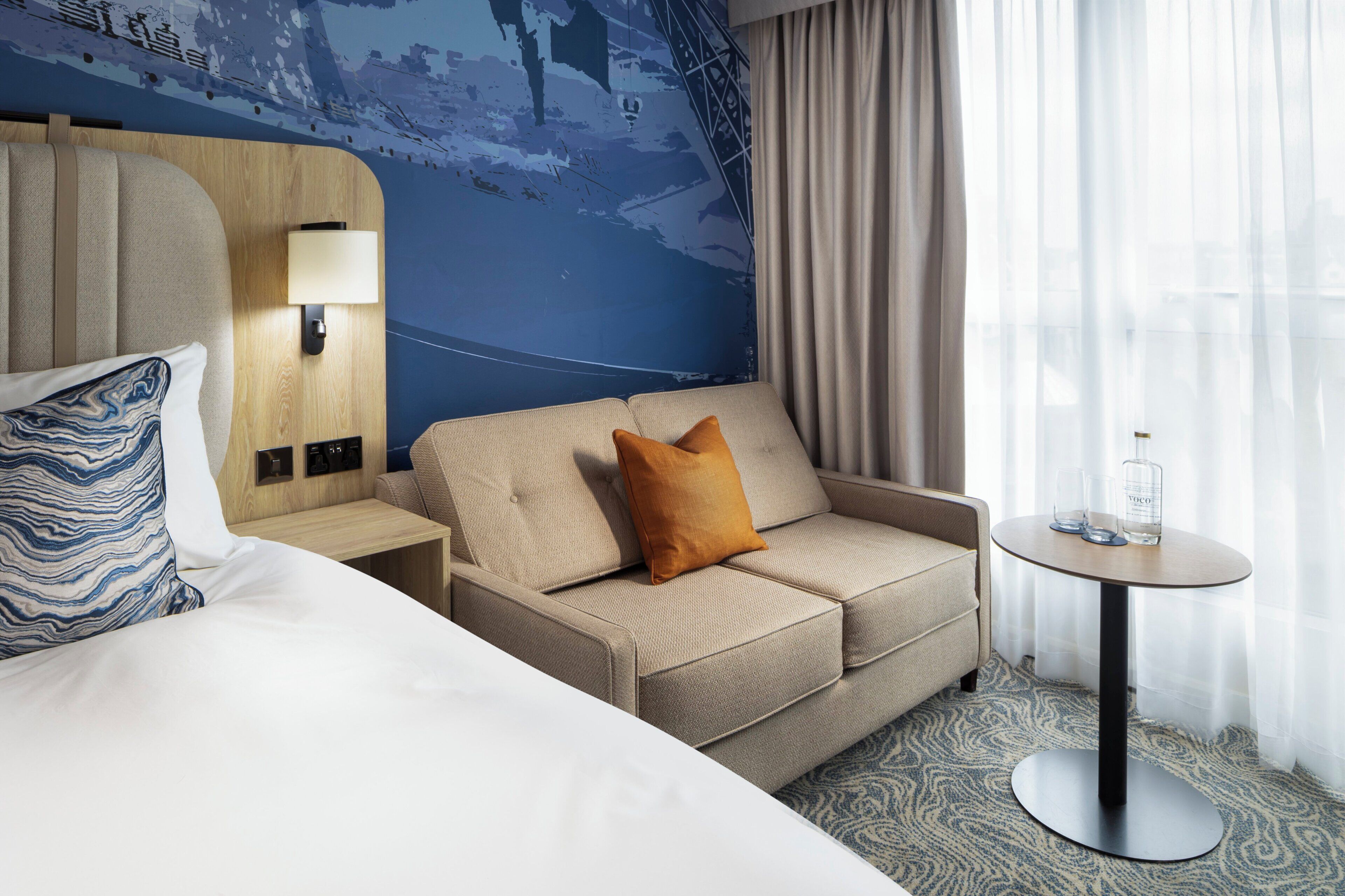 A hotel room with a bed, a sofa, a small table with a lamp, and a wall mural.