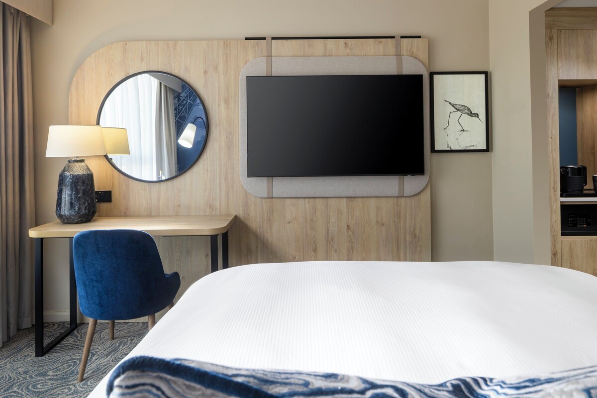 Habitació d'hotel amb un llit, un escriptori amb una làmpada, un televisor i un quadre al mur.