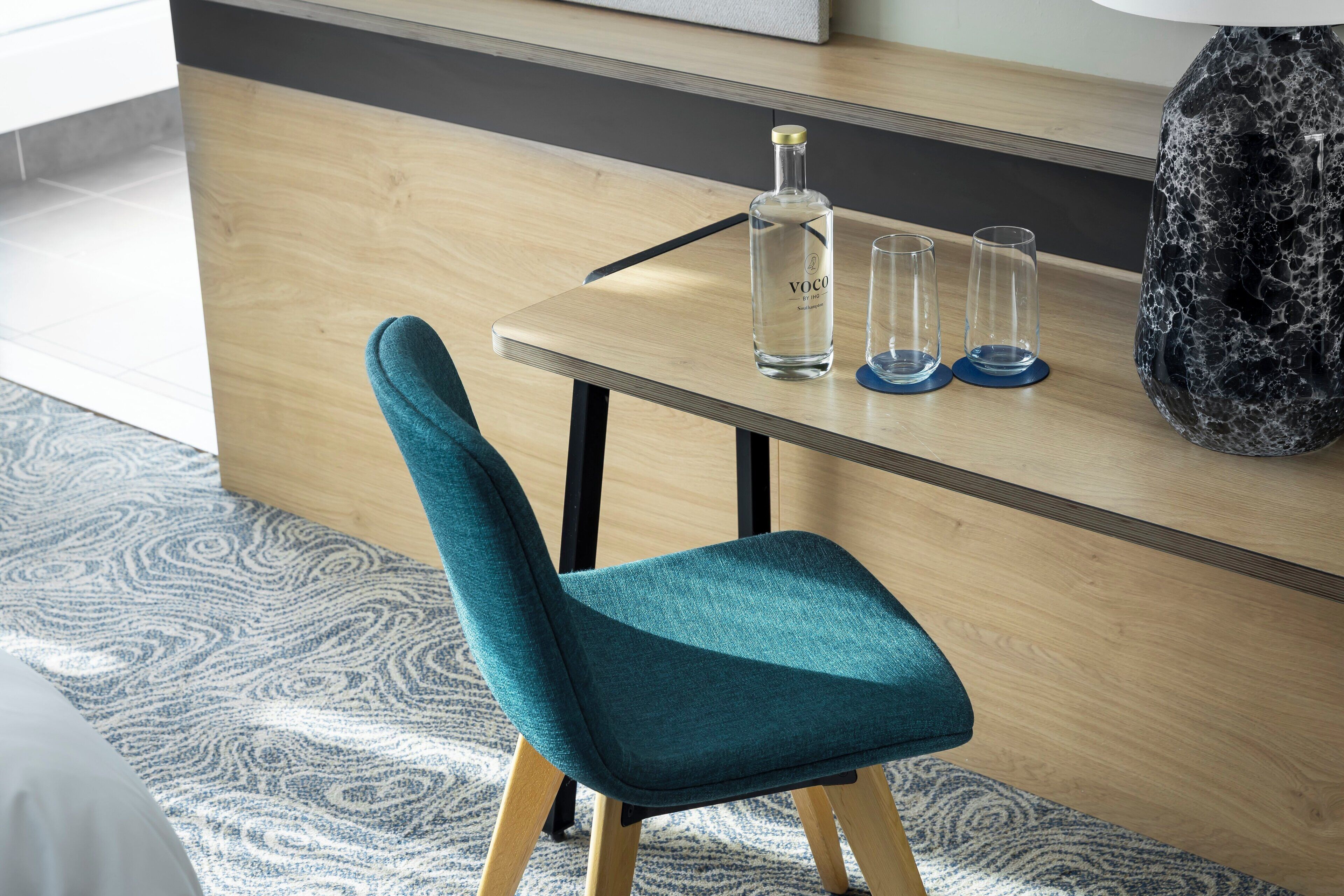 Una silla color aguamarina con patas de madera, una mesa de madera con una botella y vasos, y un florero negro.