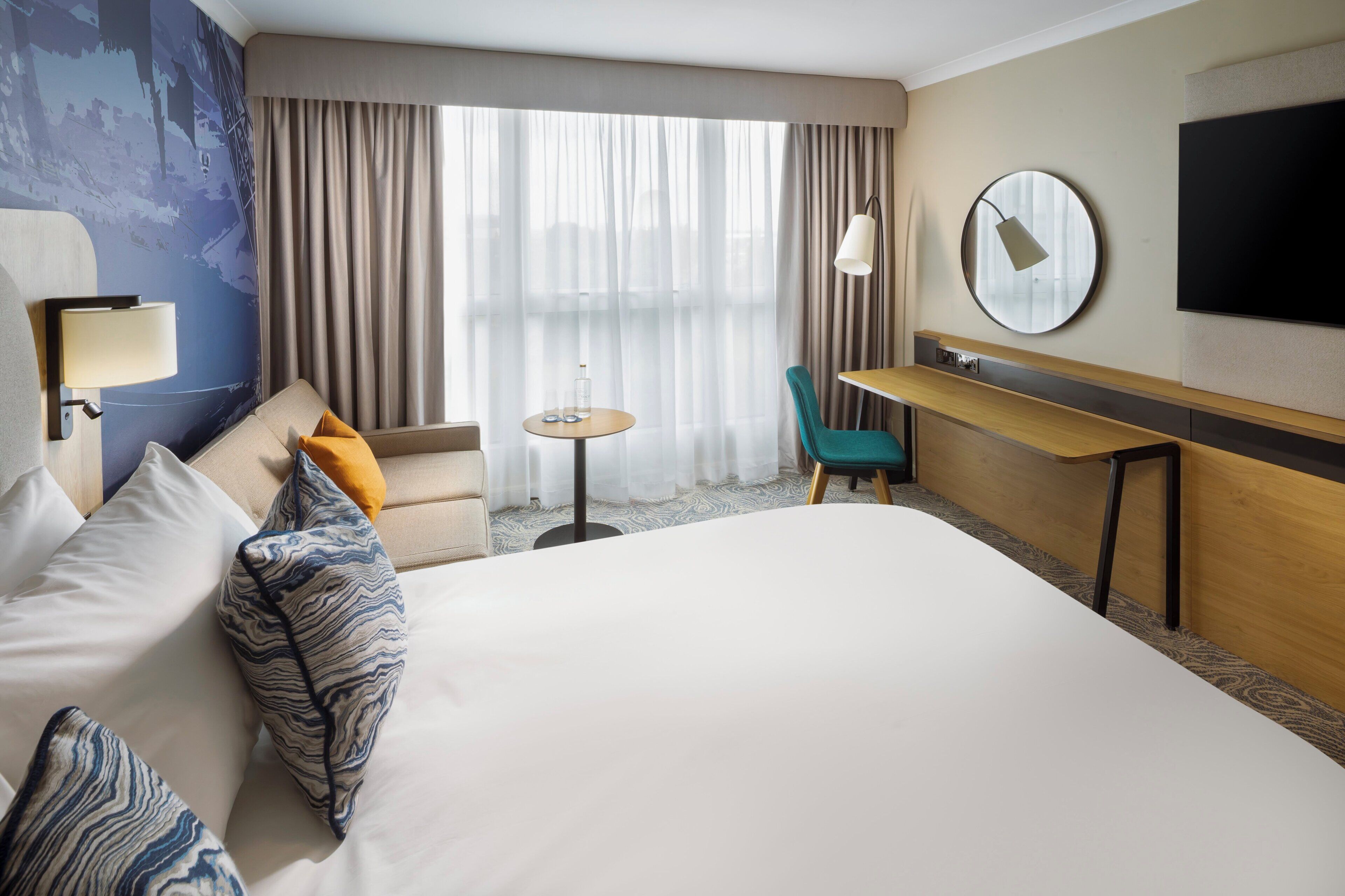 A hotel room with a large bed, a desk, a chair, a small table, a TV, and a window with curtains.