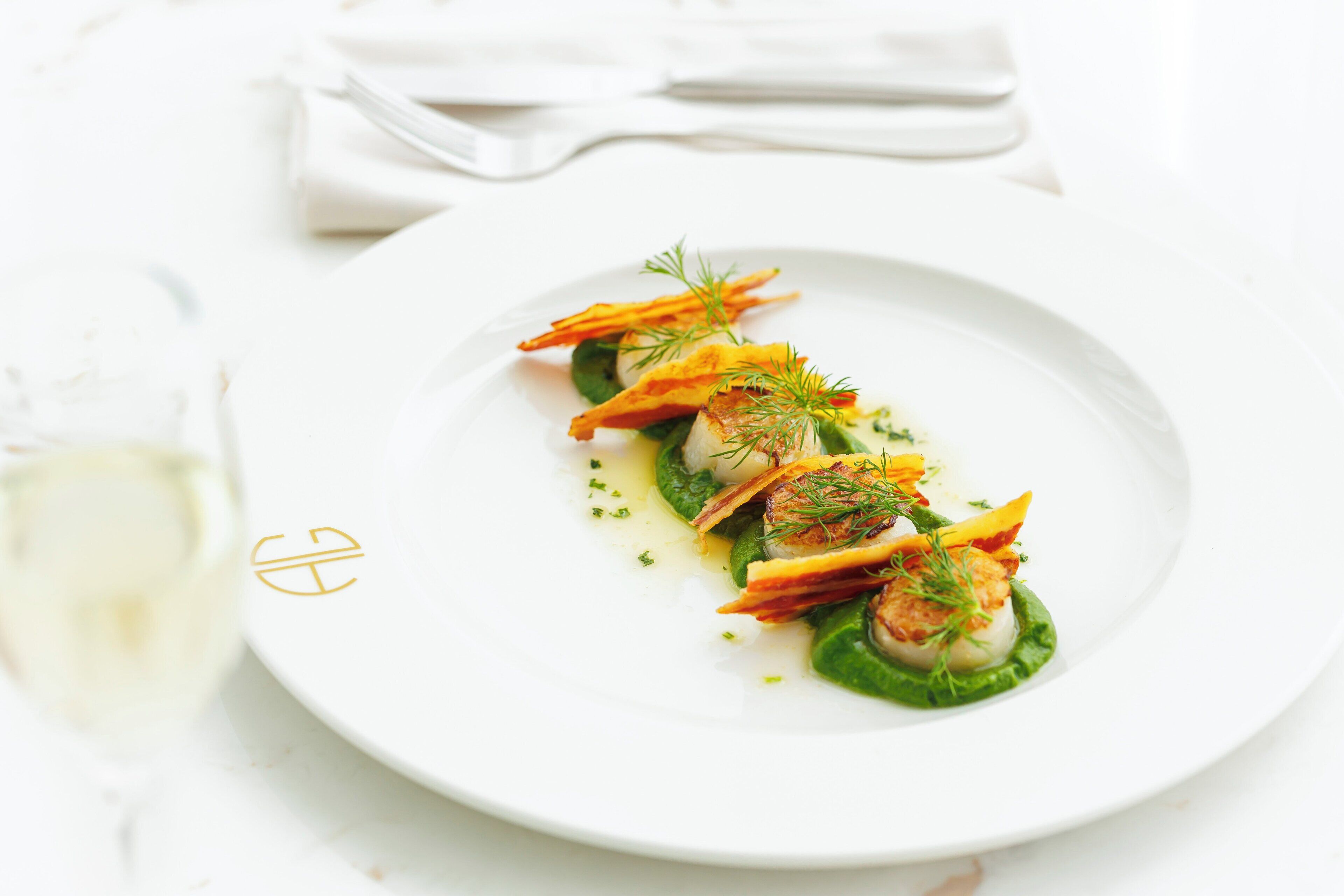 A white plate with a dish of food, a napkin, and a glass of white wine on a white surface.
