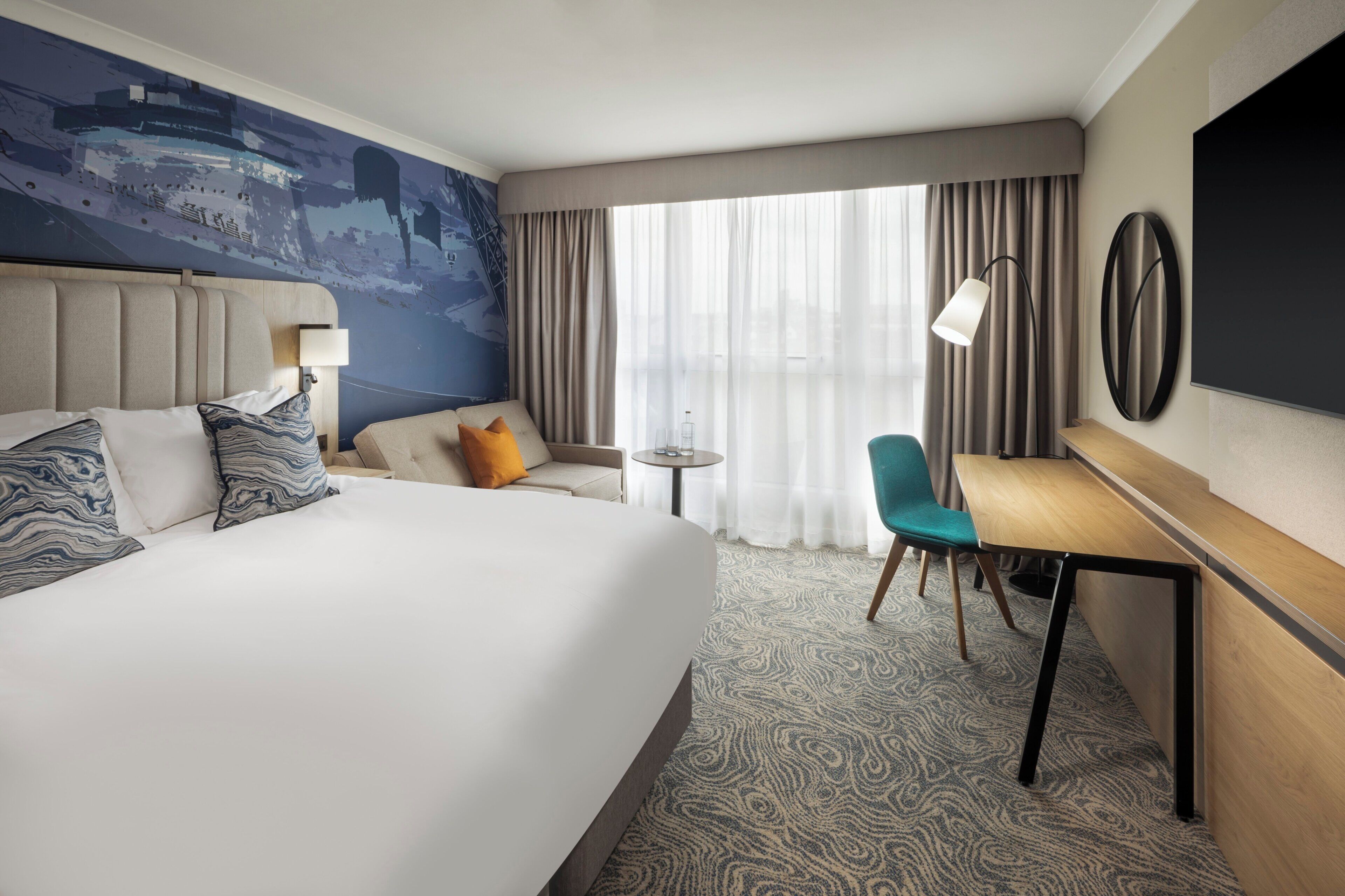 A hotel room with a large bed, a desk, a chair, a small table, and a TV.