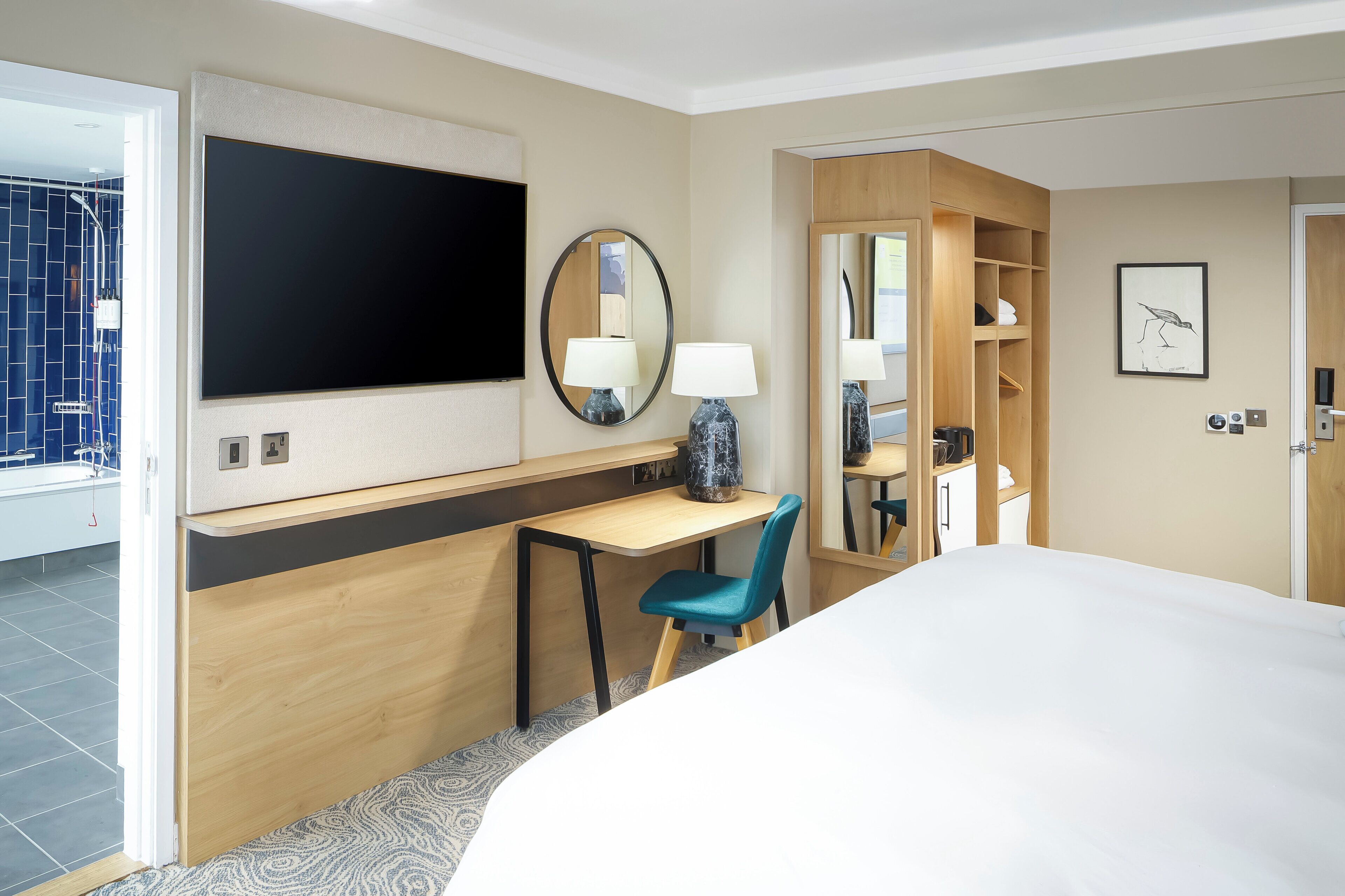 A hotel room with a bed, a desk with a chair, a TV, a mirror, a wardrobe, and a bathroom visible through an open door.