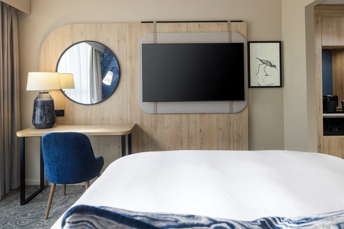 A hotel room with a bed, a desk with a lamp, a TV, and a framed picture on the wall.