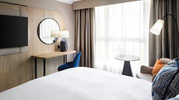 Habitación de hotel con una cama grande, un televisor montado en la pared, un espejo redondo, un escritorio con lámpara, una mesita y una silla.
