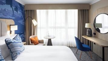 A hotel room with a large bed, a desk with a chair, a round mirror, and a window with curtains.