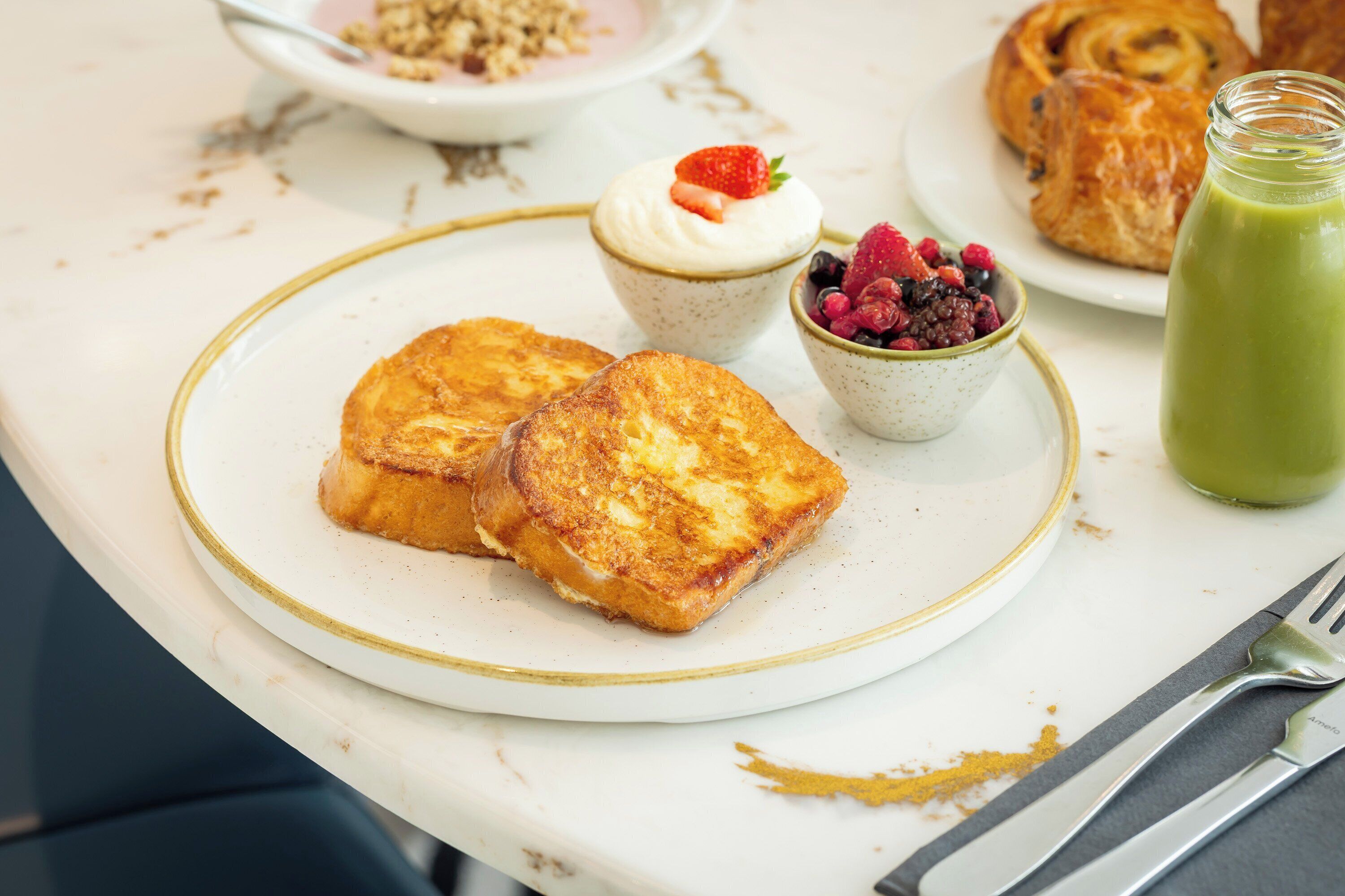 A breakfast setting with French toast, yogurt with berries, a pastry, and a green smoothie on a table.