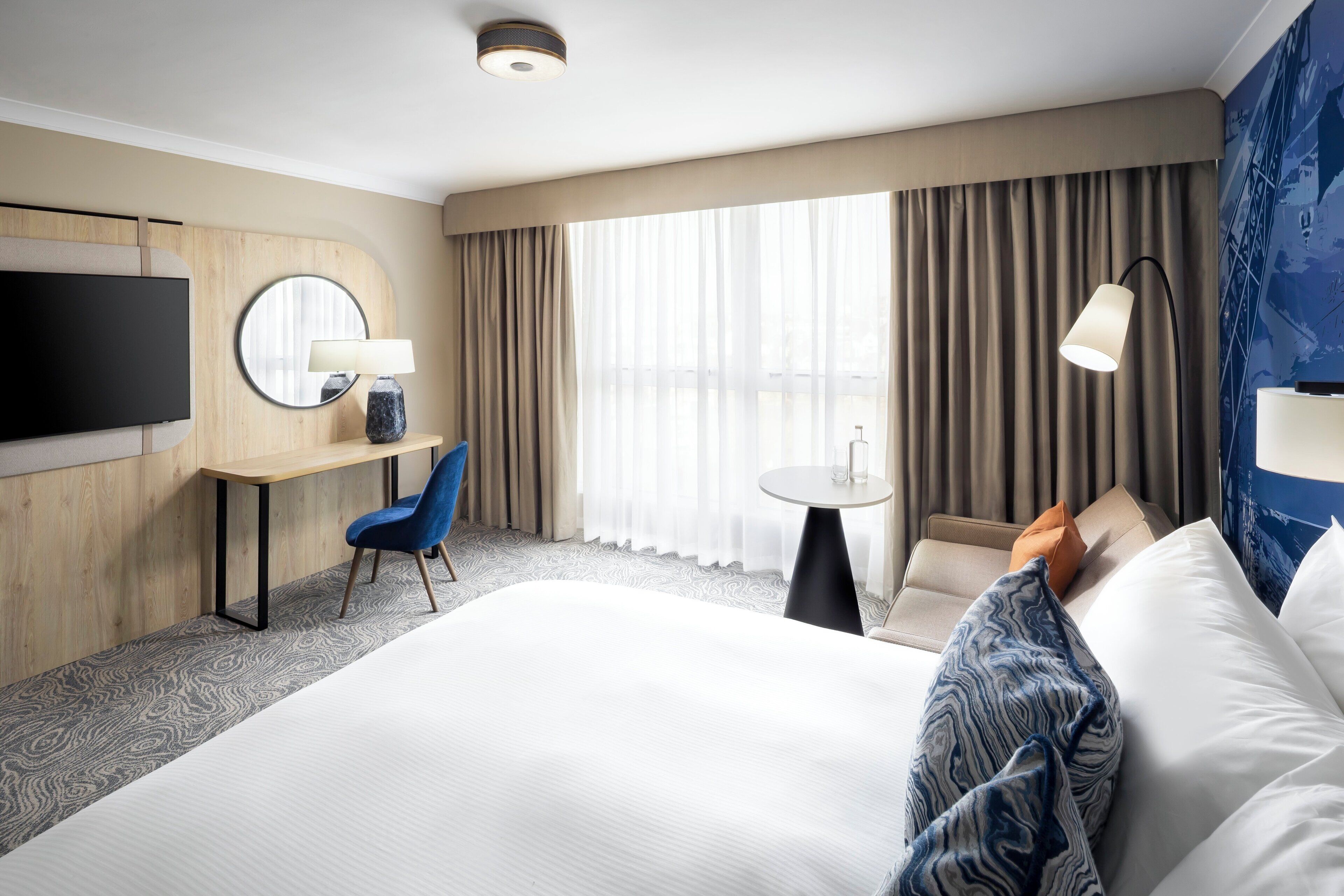 A hotel room with a bed, a desk with a chair, a TV, a lamp, and a view of the outdoors through large windows.
