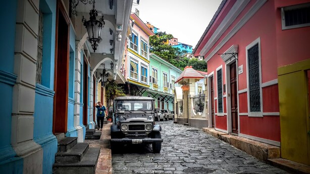 A cobblestone street lined with colorful buildings, a vintage car parked on the side, and a person walking. - Hotel Continental (Guayaquil)