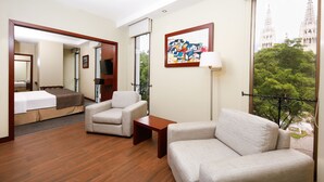A hotel room with a wooden floor, a sofa, a chair, a small table, a bed, a TV, a painting, and a balcony with a view of a cathedral. - Hotel Continental (Guayaquil)